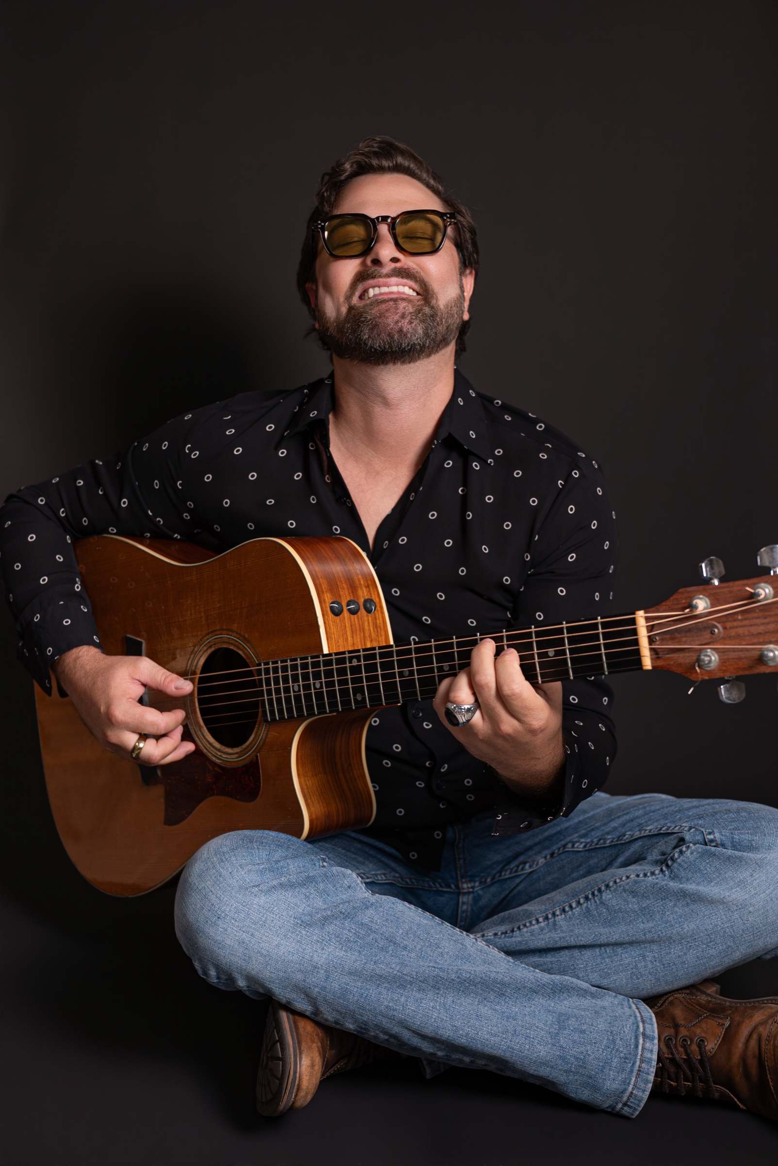 Willy B sitting cross-legged, playing an acoustic guitar and laughing with his head tilted back