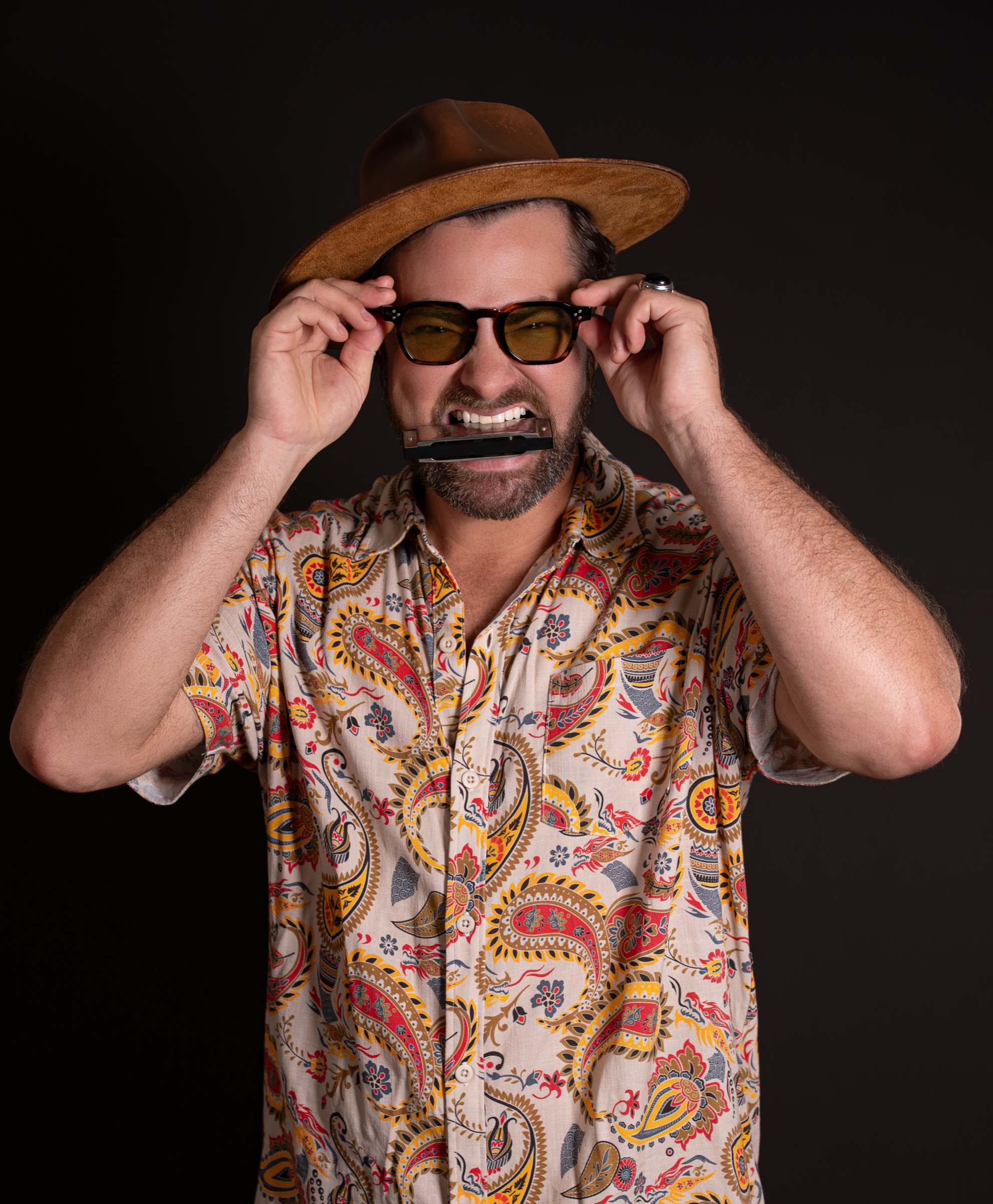 Willy B in a paisley shirt and suede hat, a harmonica between his teeth, adjusting his tinted glasses
