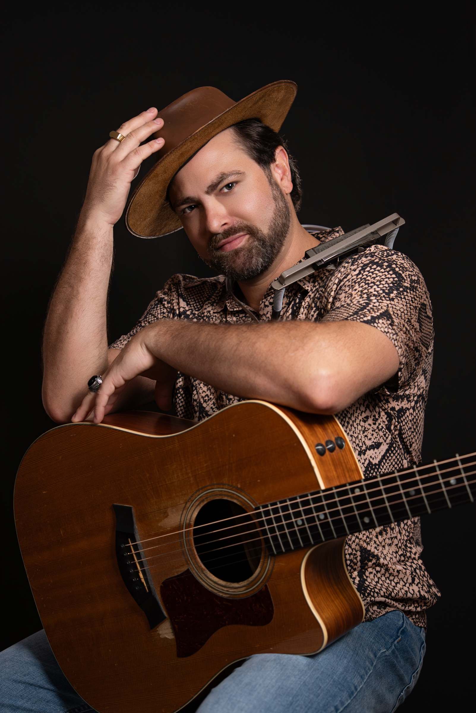 Willy B tipping a brown suede hat and smiling, a harmonica on a neck rack and an acoustic guitar in his arms
