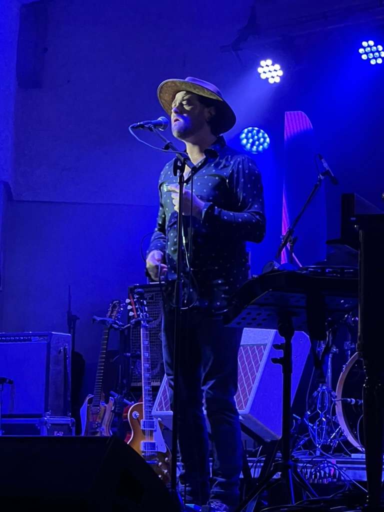 Willy B in a hat singing into a microphone on a concert stage washed in blue light, guitars on stands behind him