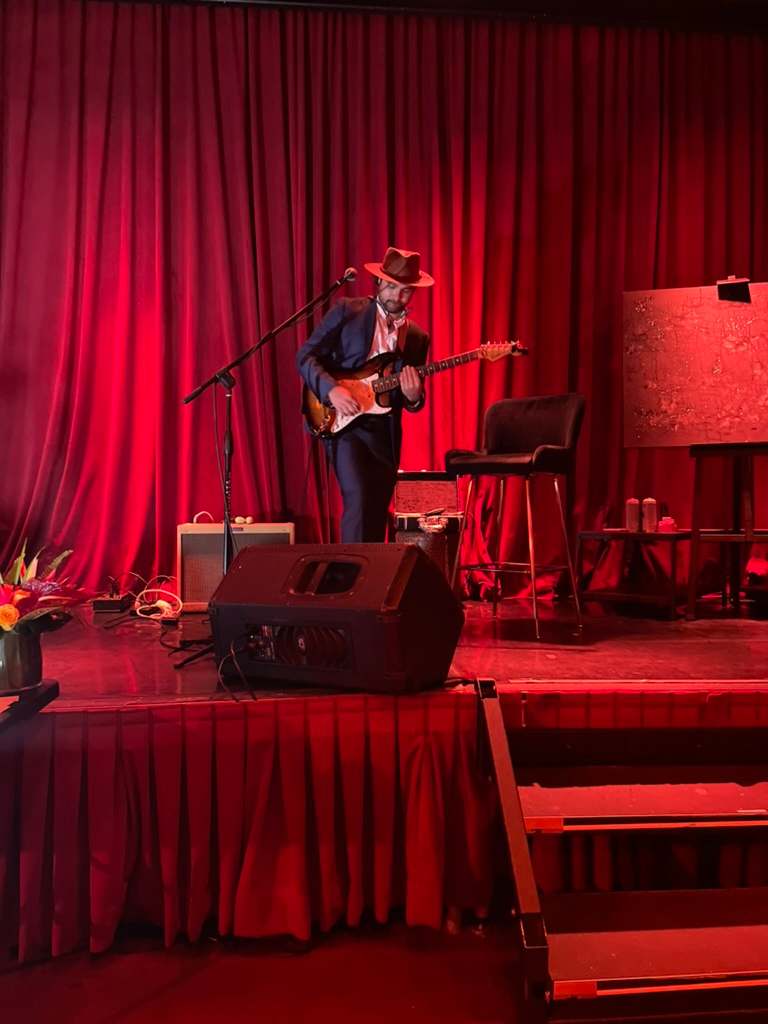 Willy B in a navy suit and brown hat playing a sunburst electric guitar on a small stage framed by red velvet curtains