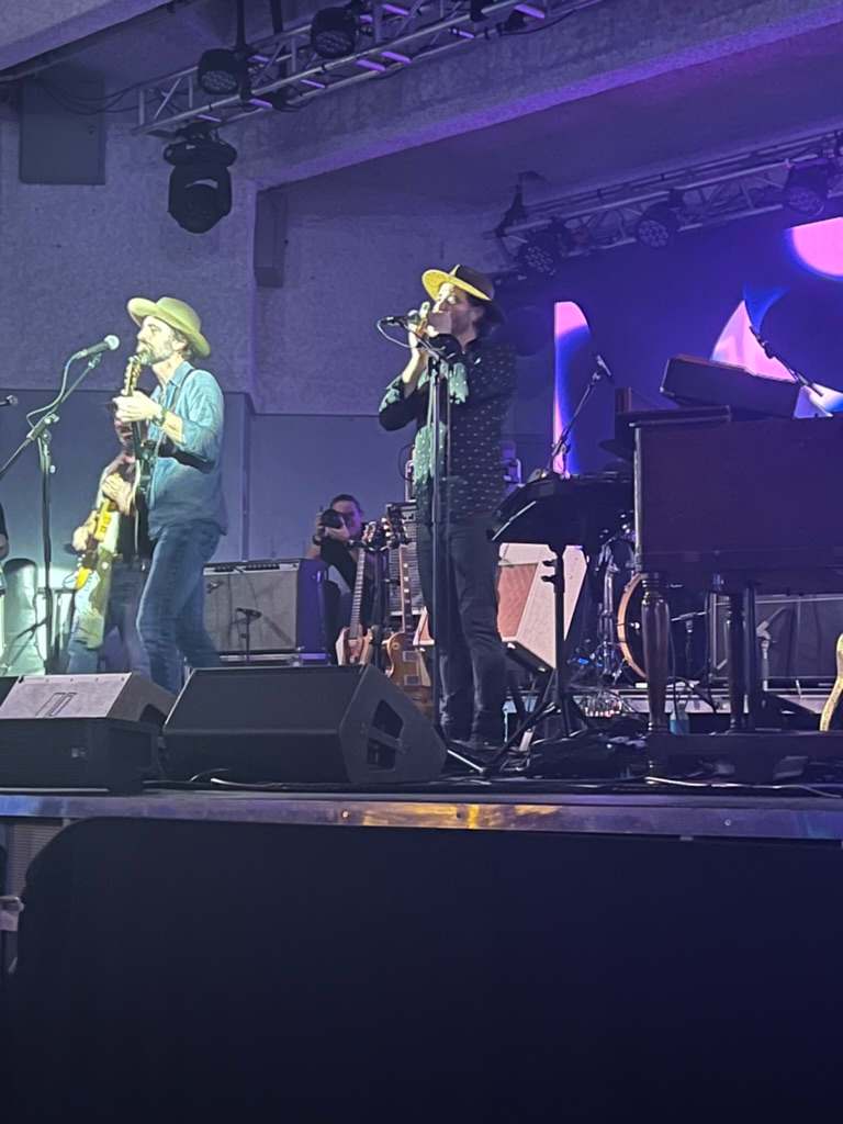 Willy B playing harmonica at the microphone on a concert stage, beside a guitarist singing at his own mic