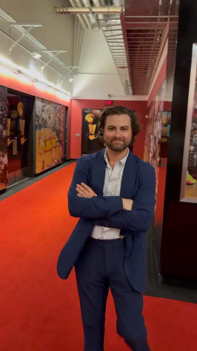 Willy B in a navy suit and open-collared shirt, arms crossed and smiling, in a red-carpeted hallway lined with framed photos