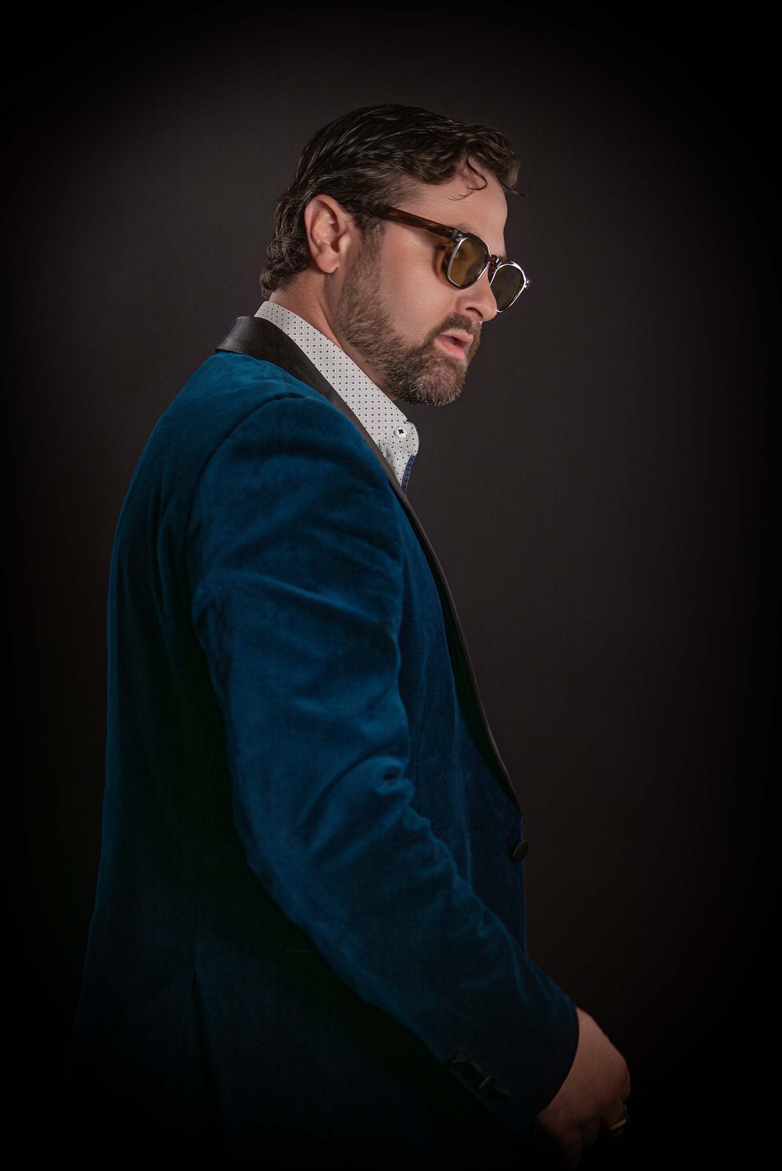 Willy B in a teal velvet blazer and tinted glasses, looking back over his shoulder against a dark backdrop